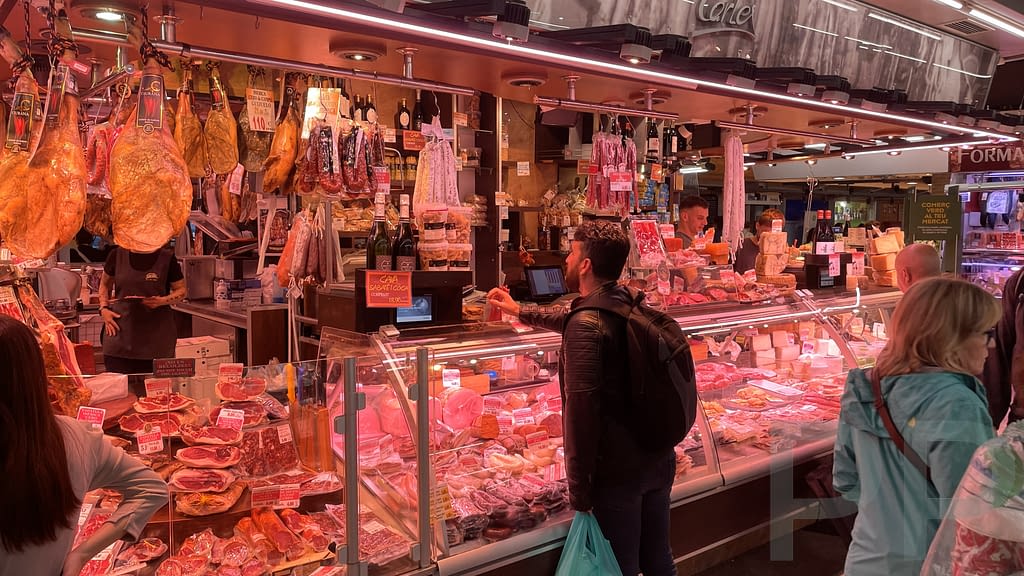 Market stall in Barcelona displaying cured meats and cheeses as shoppers browse and interact with vendors.
