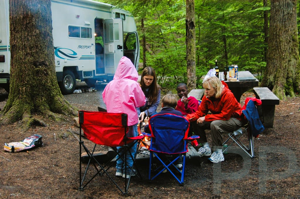 Family sitting around a campfire at a forest campsite with a Class C RV behind.