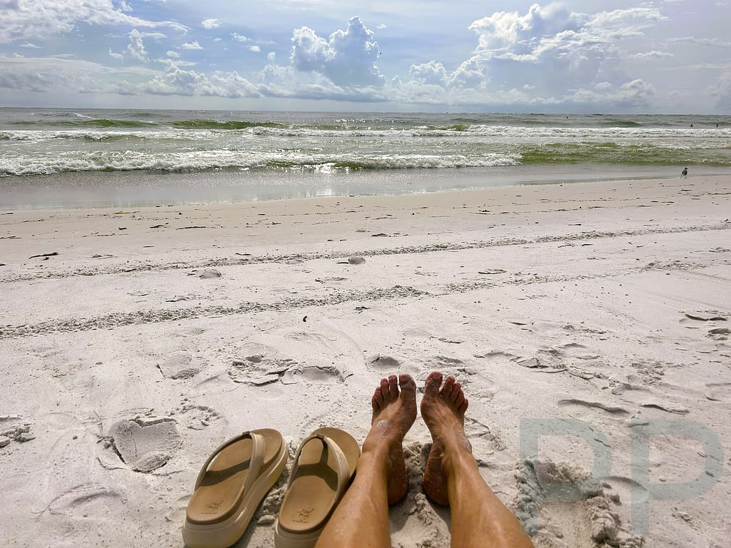 Person relaxing on white sand beach with sandals in foreground and waves beyond.