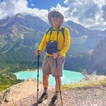 Hiker with trekking poles standing on a rocky trail overlooking turquoise alpine lake in Glacier National Park