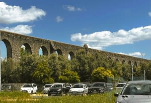 Public parking lot beneath the towering arches of &Eacute;vora&rsquo;s historic aqueduct under a blue sky.