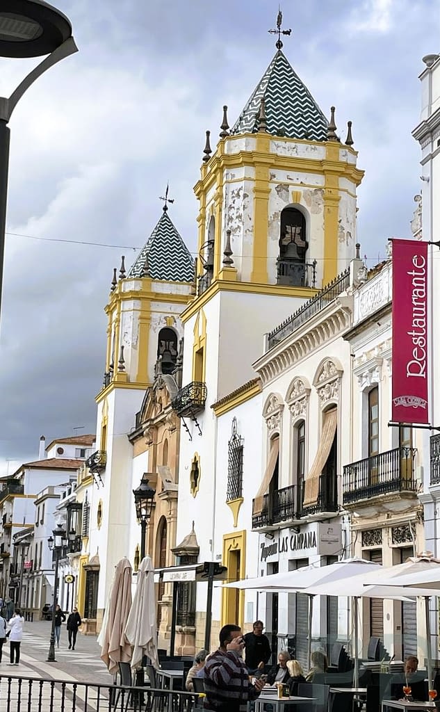 White and yellow church towers with patterned tile roofs above a busy street of cafés and shops in Ronda, Spain.