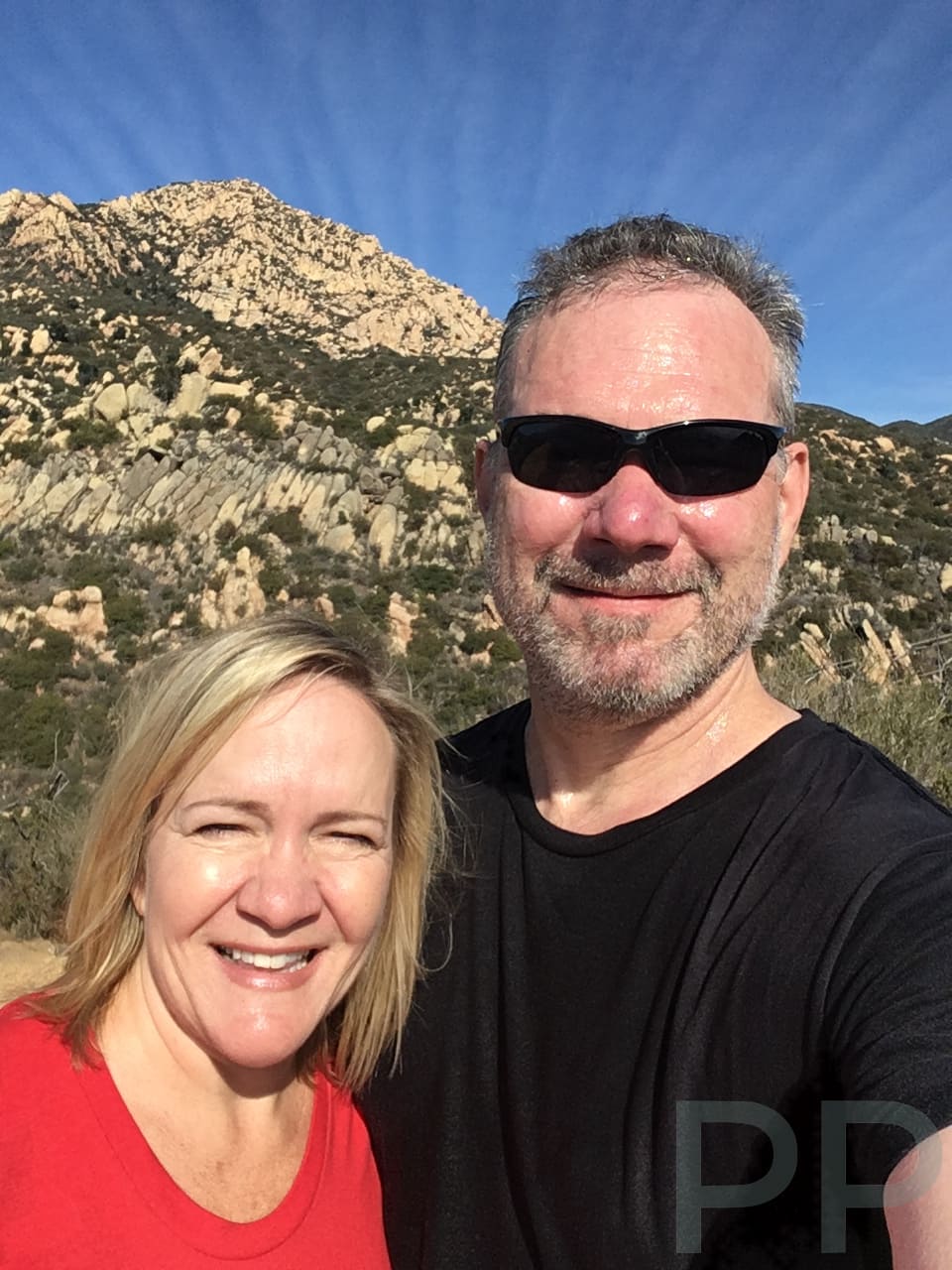 Two people smiling in a hiking selfie with a hazy mountain landscape behind them.