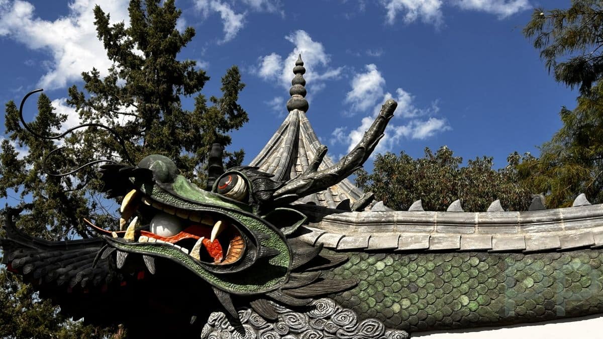 A detailed Chinese-style dragon sculpture arching over a white moon-gate entrance at Botanica Wichita, with scales, claws, and expressive features highlighted against trees and blue sky.