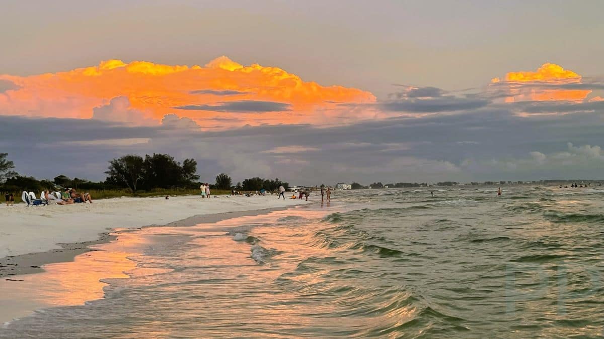People walking and relaxing on the beach at sunset on Anna Maria Island, with glowing orange clouds reflecting on the Gulf waves.