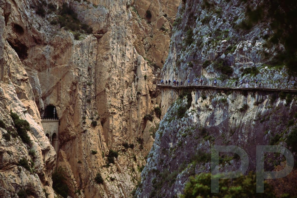 Hikers on the Caminito del Rey boardwalk clinging to a vertical cliff, with a train tunnel entrance visible in the gorge wall.