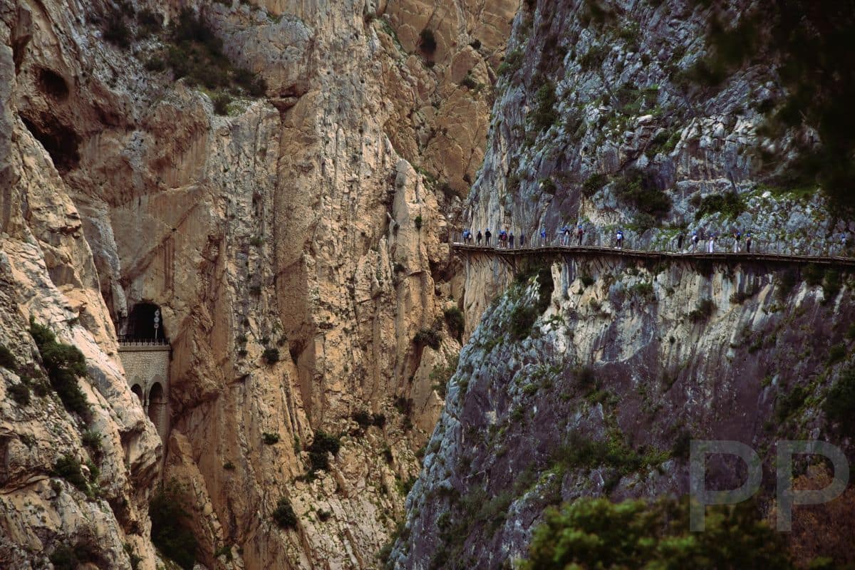 Hikers on the Caminito del Rey boardwalk clinging to a vertical cliff, with a train tunnel entrance visible in the gorge wall.