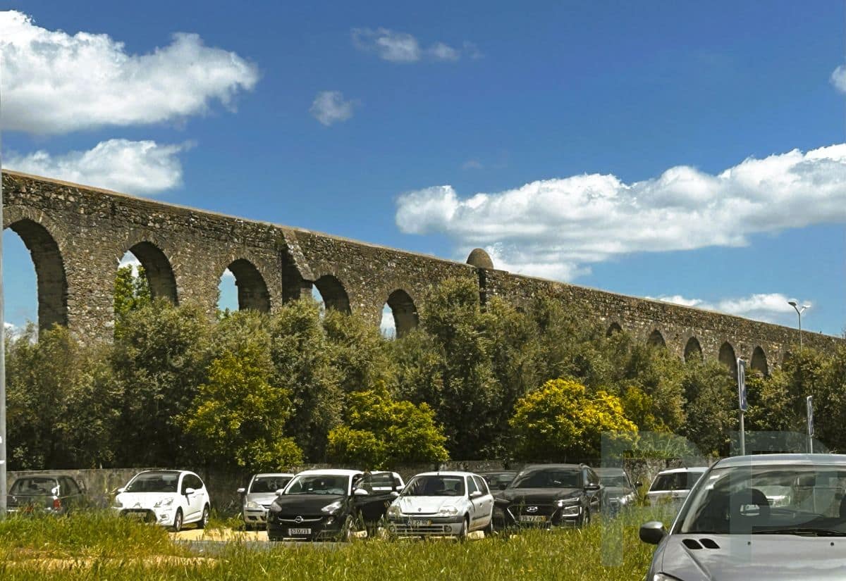 Public parking lot beneath the towering arches of Évora’s historic aqueduct under a blue sky.