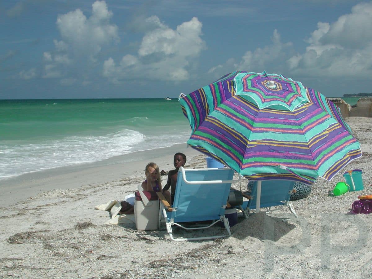 Children sitting under a colorful striped umbrella on the beach with beach chairs, cooler, and sand toys at Anna Maria Island.