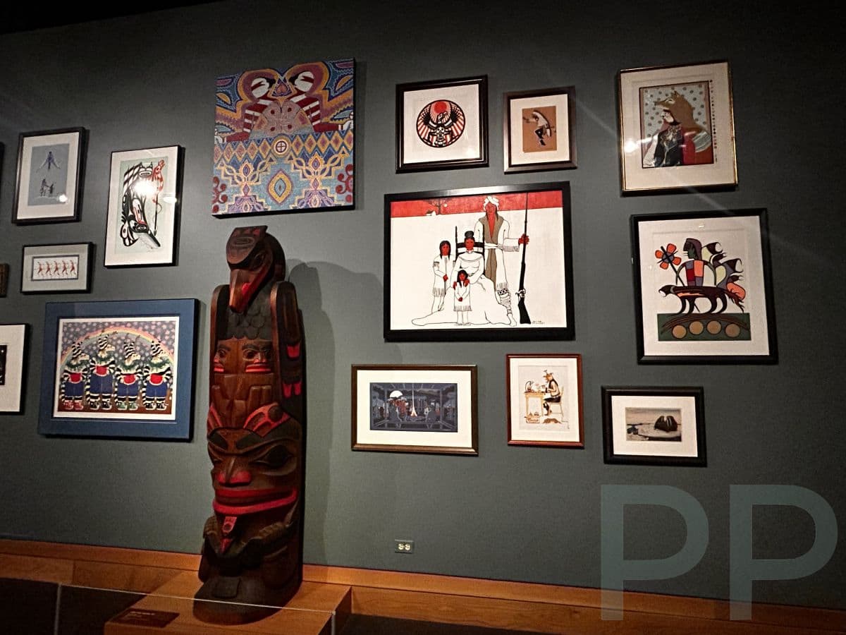 Gallery wall at the Wichita Art Museum featuring a collection of framed Native and contemporary artworks displayed alongside a carved totem-style sculpture.