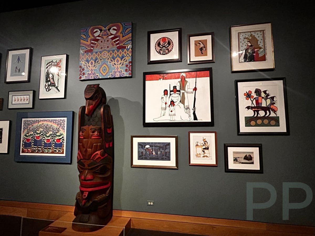 Gallery wall at the Wichita Art Museum featuring a collection of framed Native and contemporary artworks displayed alongside a carved totem-style sculpture.