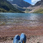 Hiking shoes resting on a rocky lakeshore with clear water and mountain peaks in the background.