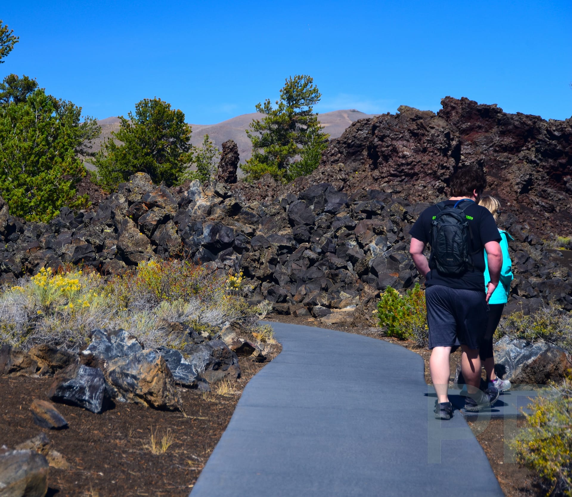 Visitors hiking a paved trail through dark volcanic cinders on the slope of a cinder cone