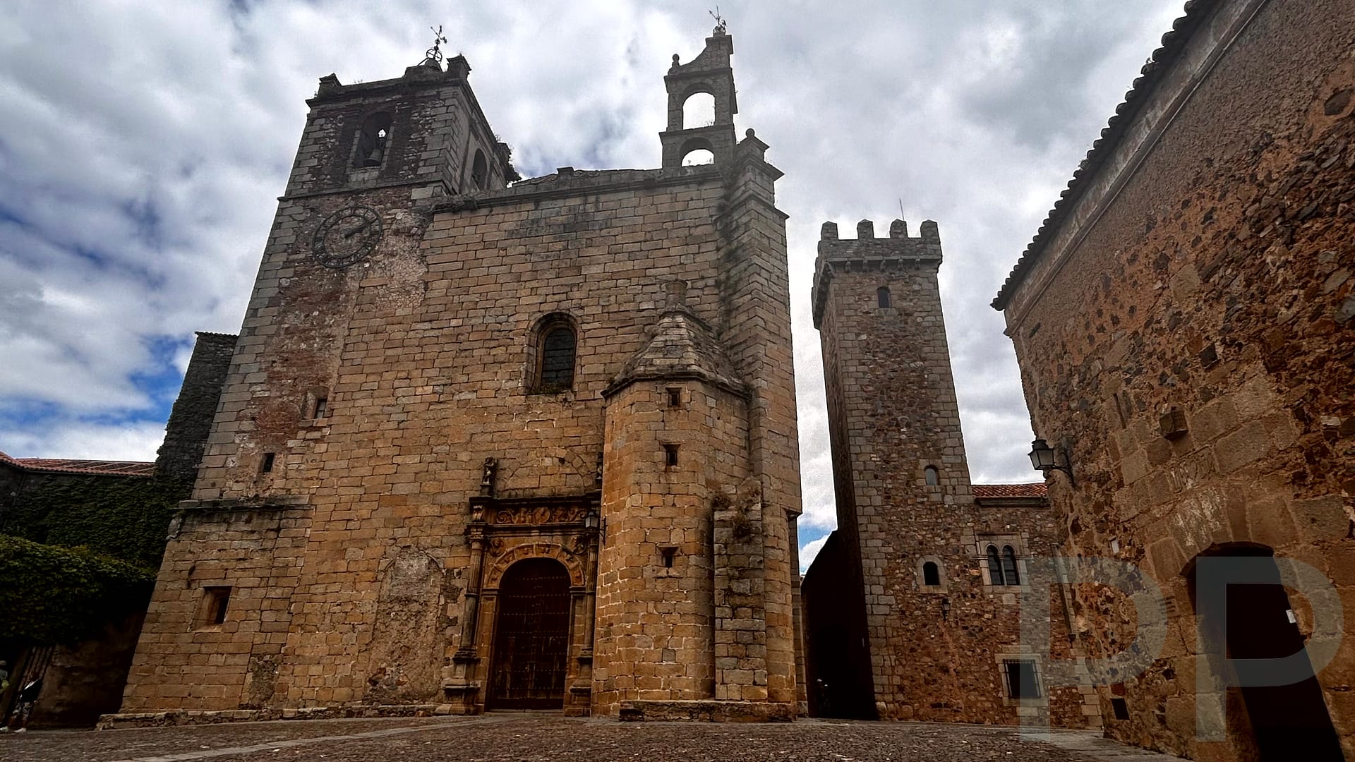 View of Plaza de Santa María in Cáceres, Spain, featuring historic buildings and the towering Concatedral de Santa María under a cloudy sky.