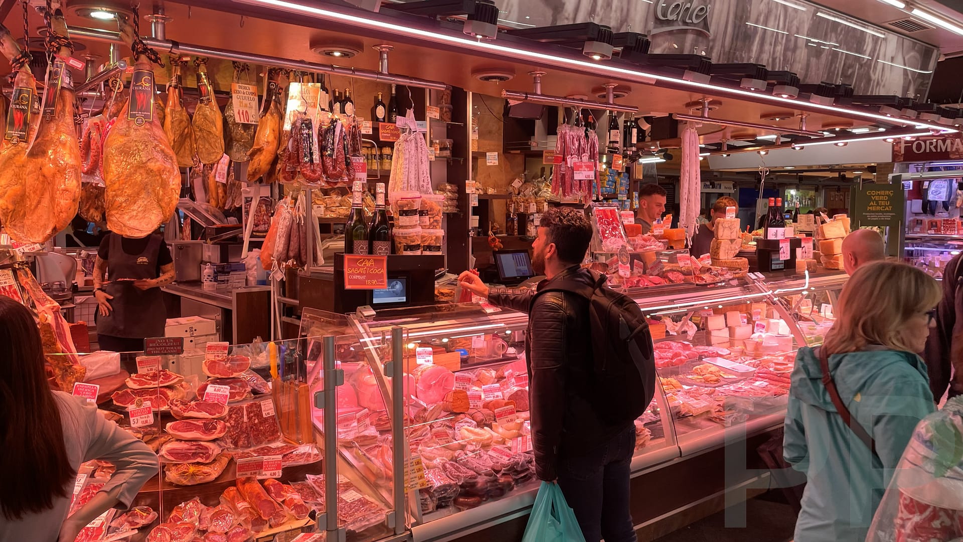 Market stall in Barcelona displaying cured meats and cheeses as shoppers browse and interact with vendors.