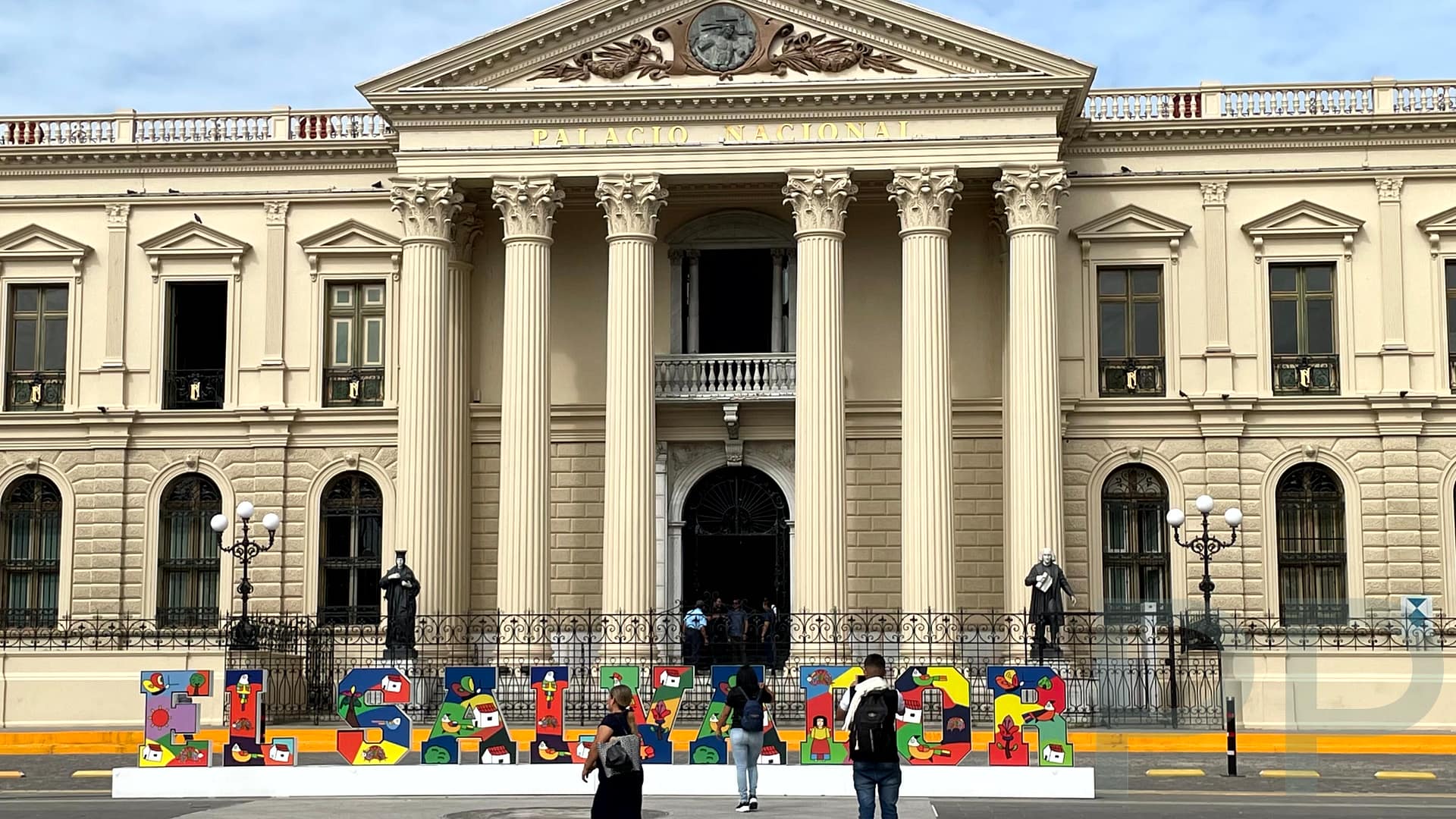 Front view of El Salvador’s National Palace in San Salvador with tall columns, statues, and colorful “El Salvador” letters in the plaza.