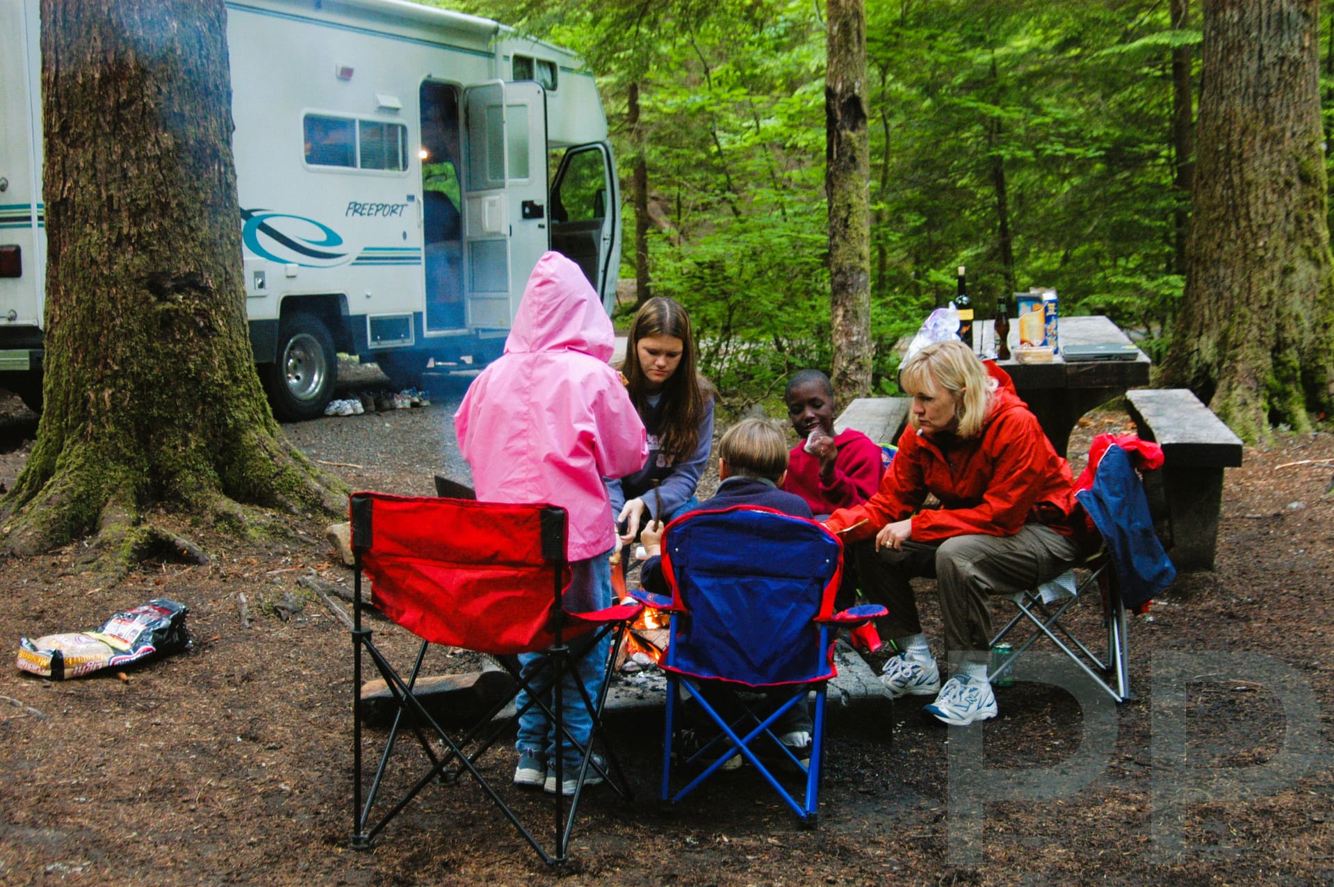 Family sitting around a campfire at a forest campsite with a Class C RV behind.