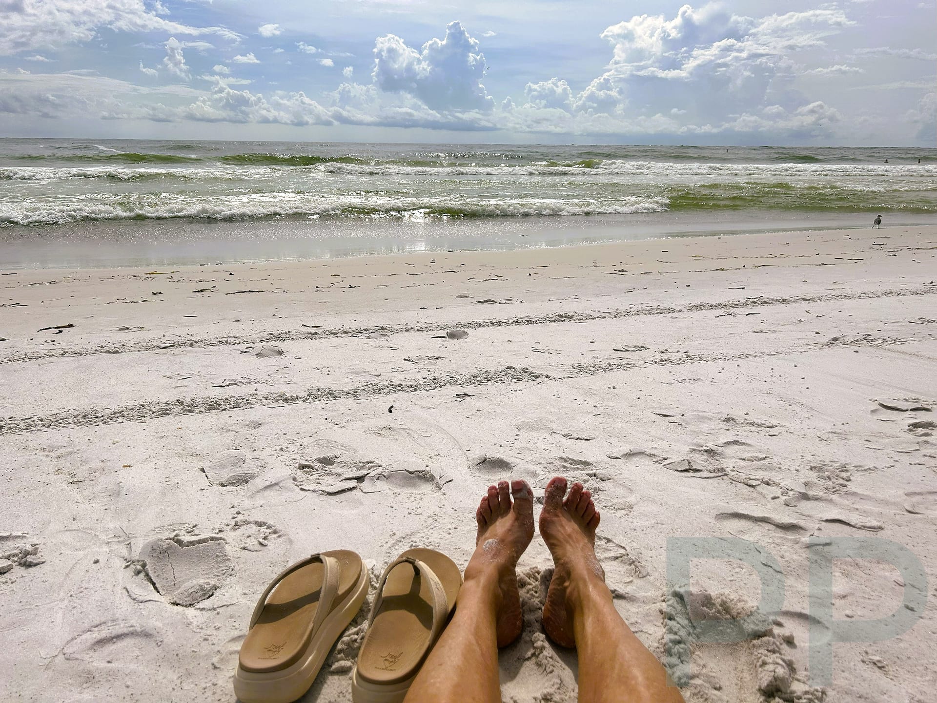 Person relaxing on white sand beach with sandals in foreground and waves beyond.