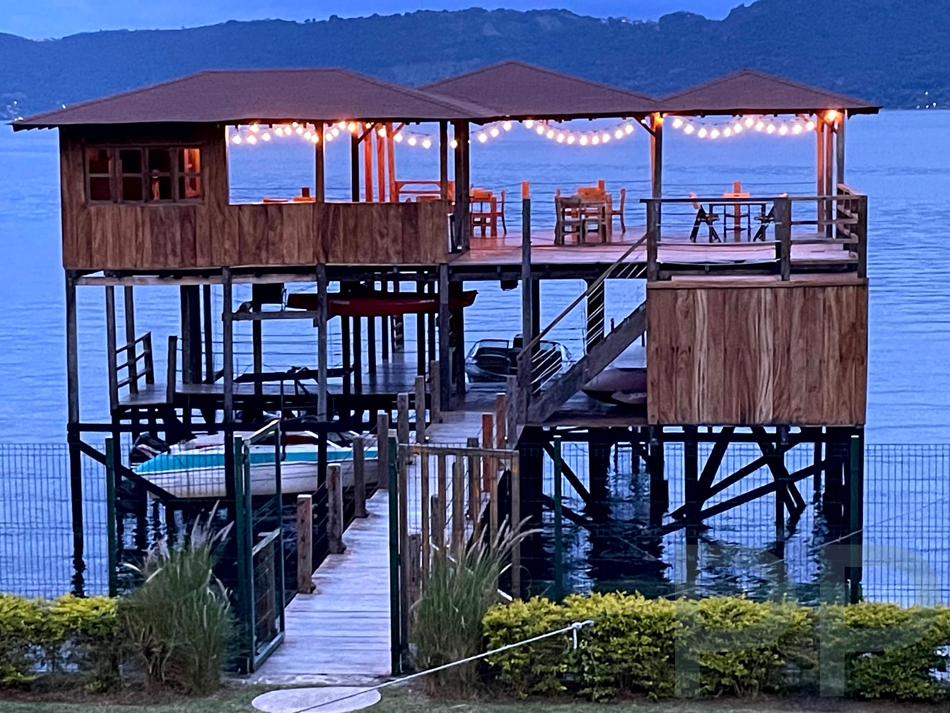 Wooden over-water deck with string lights and tables above the calm blue surface of Lago de Coatepeque, El Salvador.
