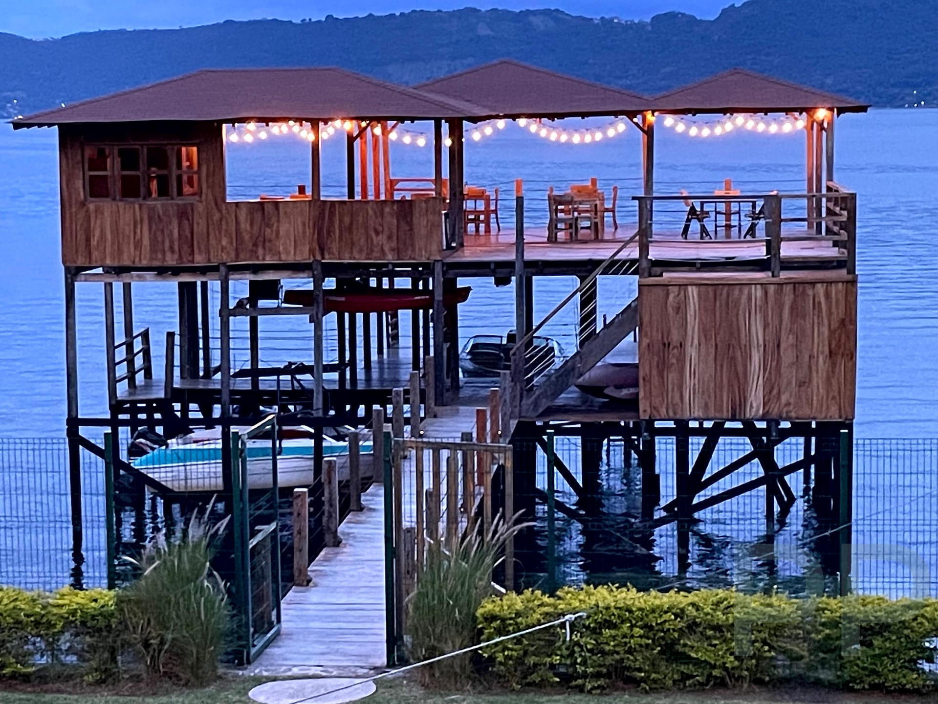 Wooden over-water deck with string lights and tables above the calm blue surface of Lago de Coatepeque, El Salvador.
