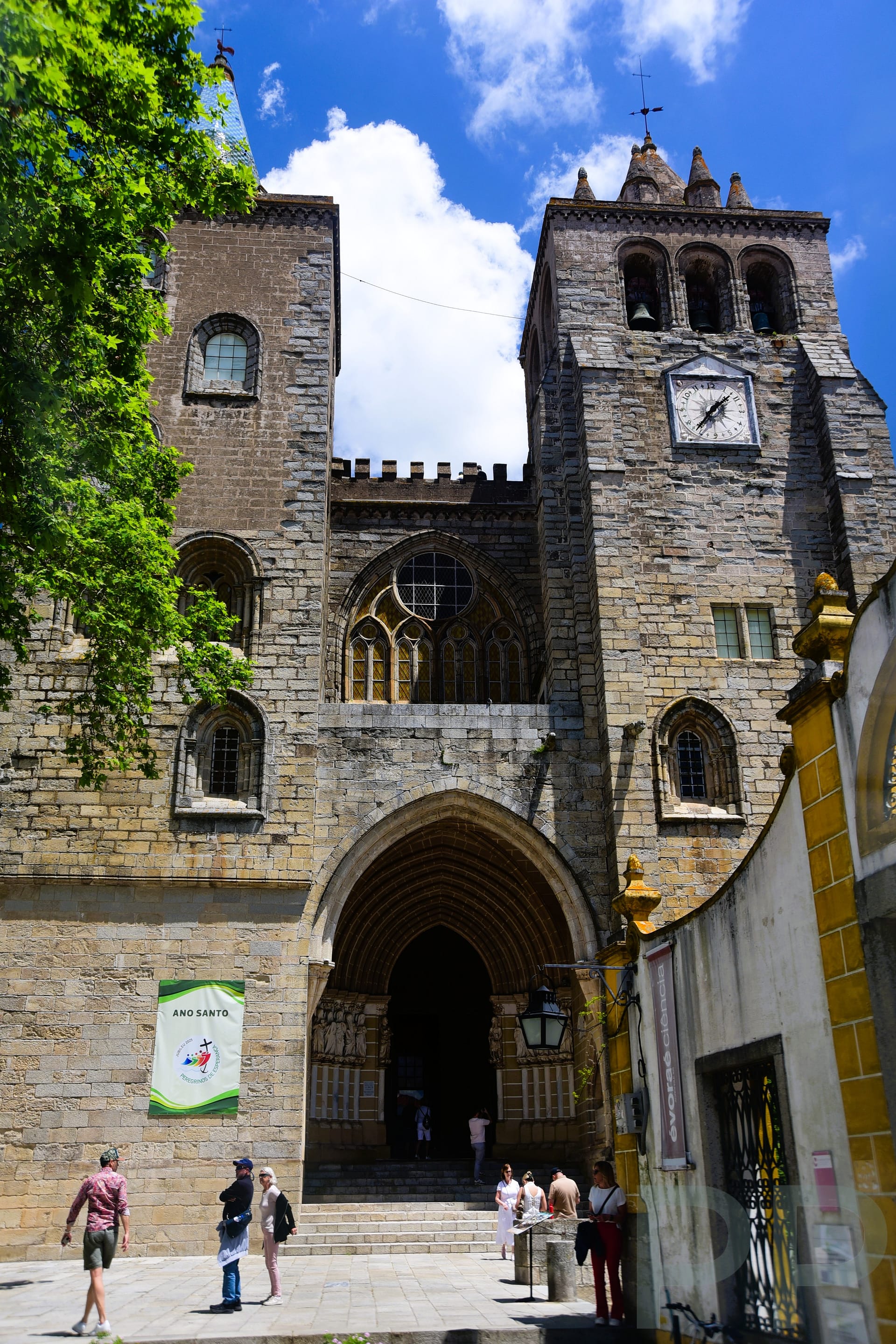 Front view of Évora Cathedral with twin stone towers, arched entrance, and clock tower against a bright blue sky.