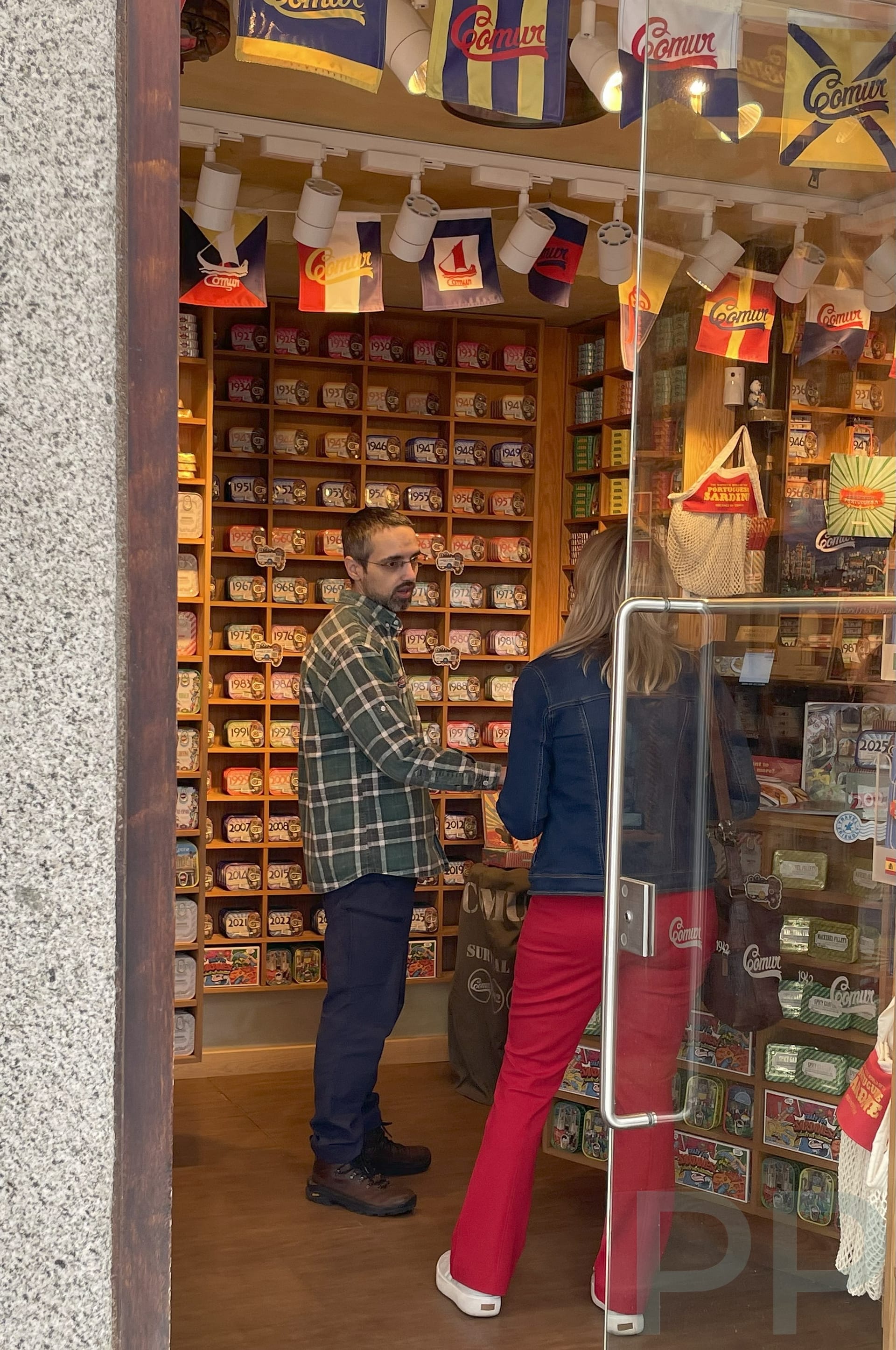 Interior of O Mundo Fantástico da Sardinha Portuguesa shop in Évora, with colorful tins of sardines stacked on the wall and a staff member explaining options to a visitor.