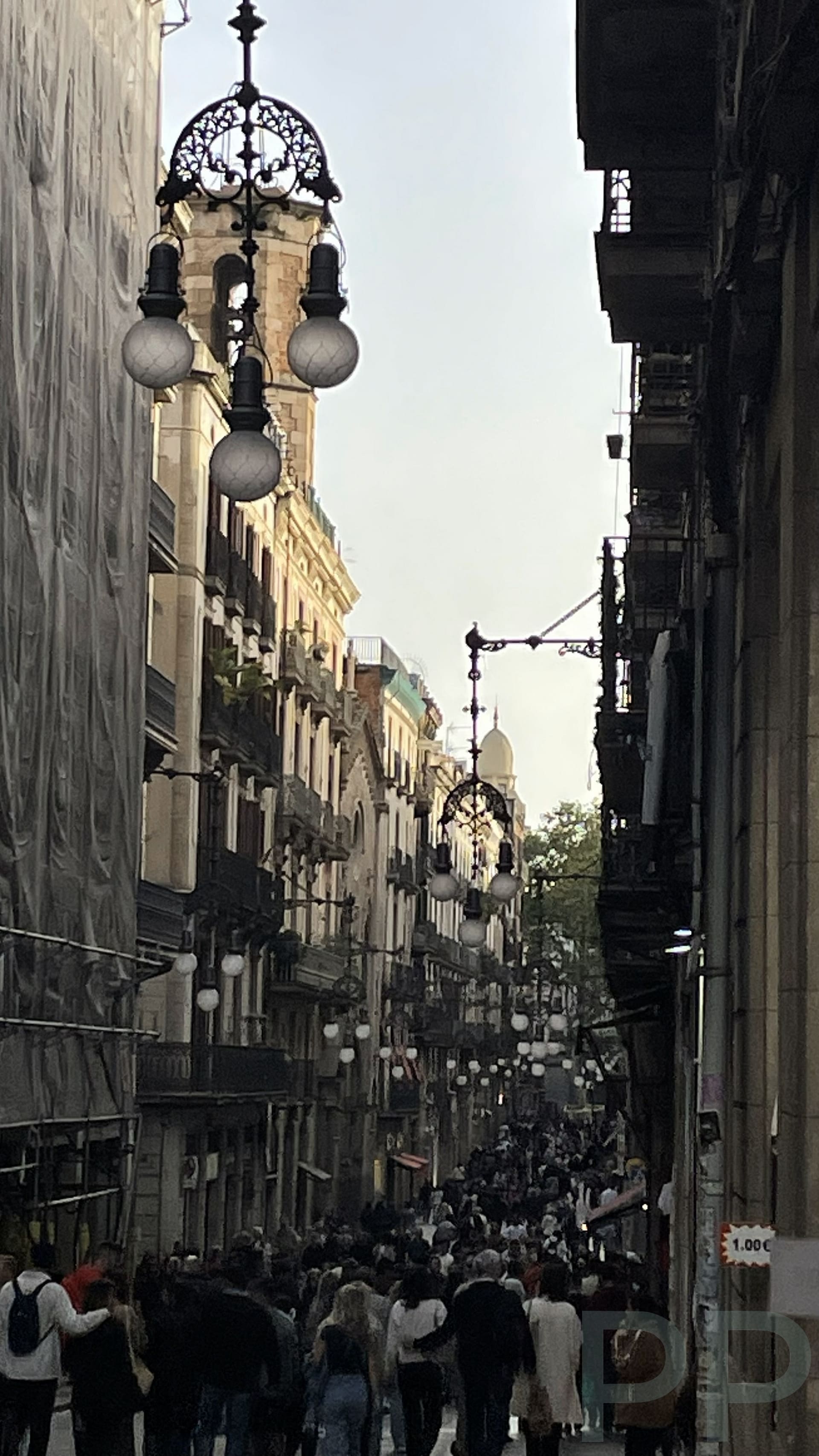 Crowded pedestrian street in Barcelona&rsquo;s old city during the early evening, with people strolling beneath historic buildings.