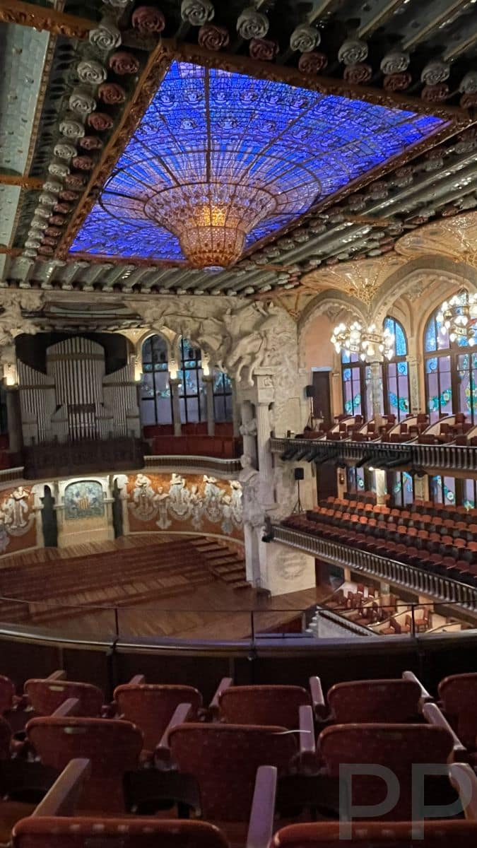 Interior of Palau de la M&uacute;sica Catalana concert hall in Barcelona, featuring the colorful stained-glass skylight, ornate balconies, and sculpted stage.