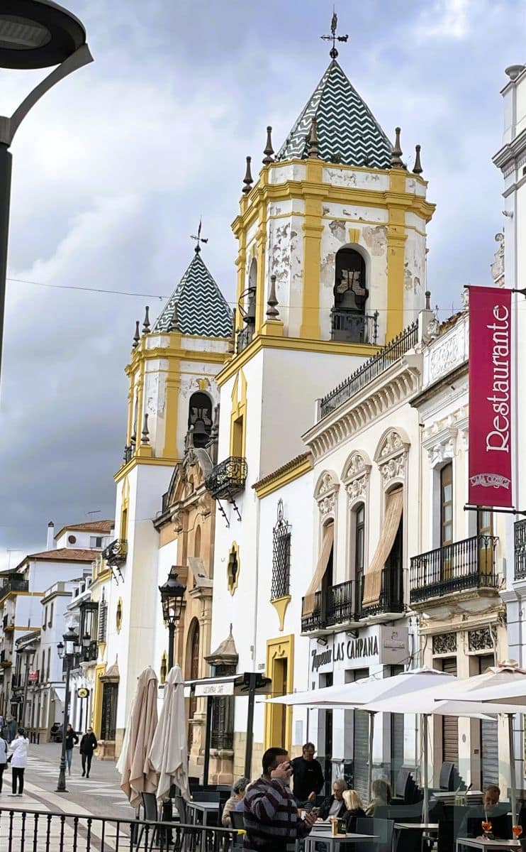 White and yellow church towers with patterned tile roofs above a busy street of cafés and shops in Ronda, Spain.