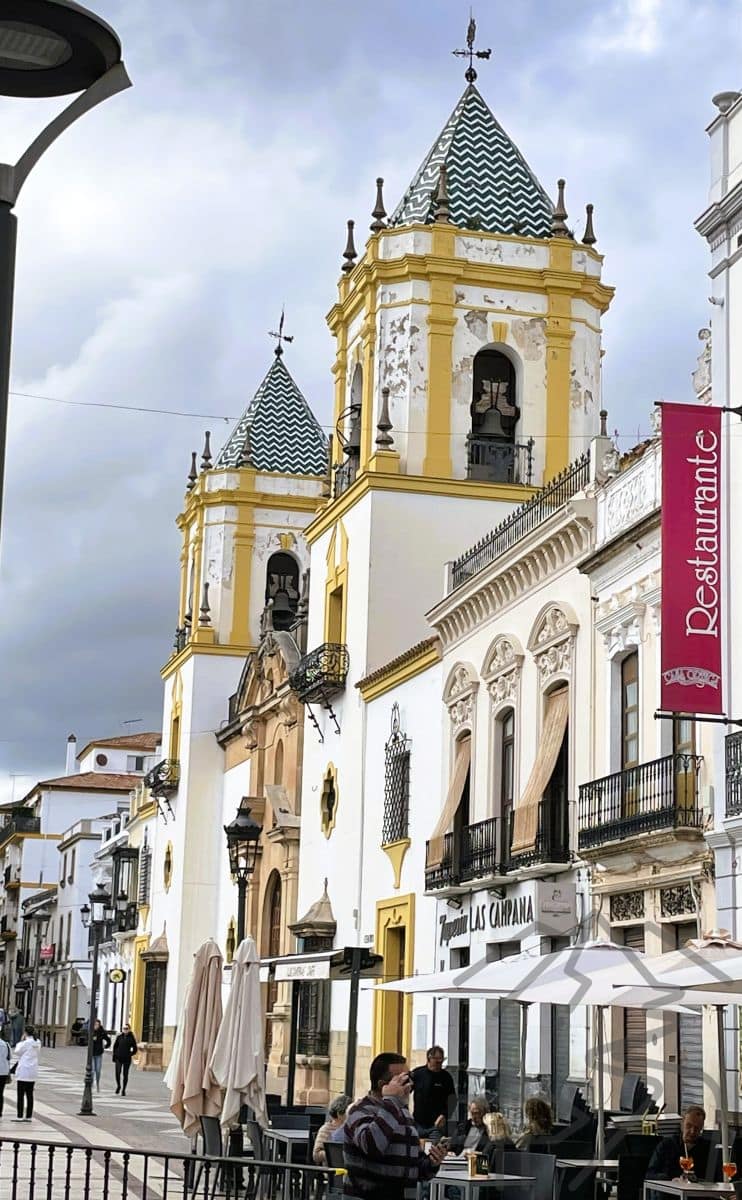 White and yellow church towers with patterned tile roofs above a busy street of caf&eacute;s and shops in Ronda, Spain.