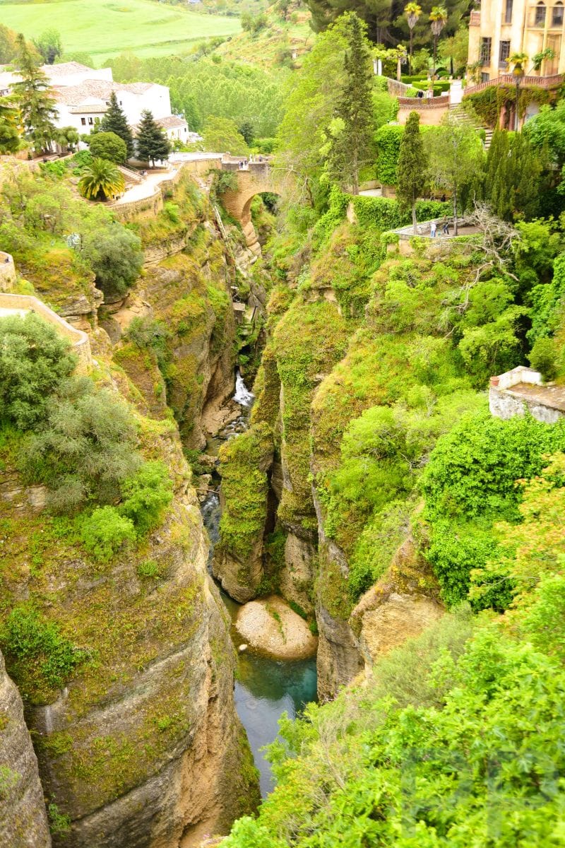 Vertical view of El Tajo Gorge in Ronda, Spain, showing steep cliffs, green vegetation, and the Guadalevín River flowing below with a small waterfall.