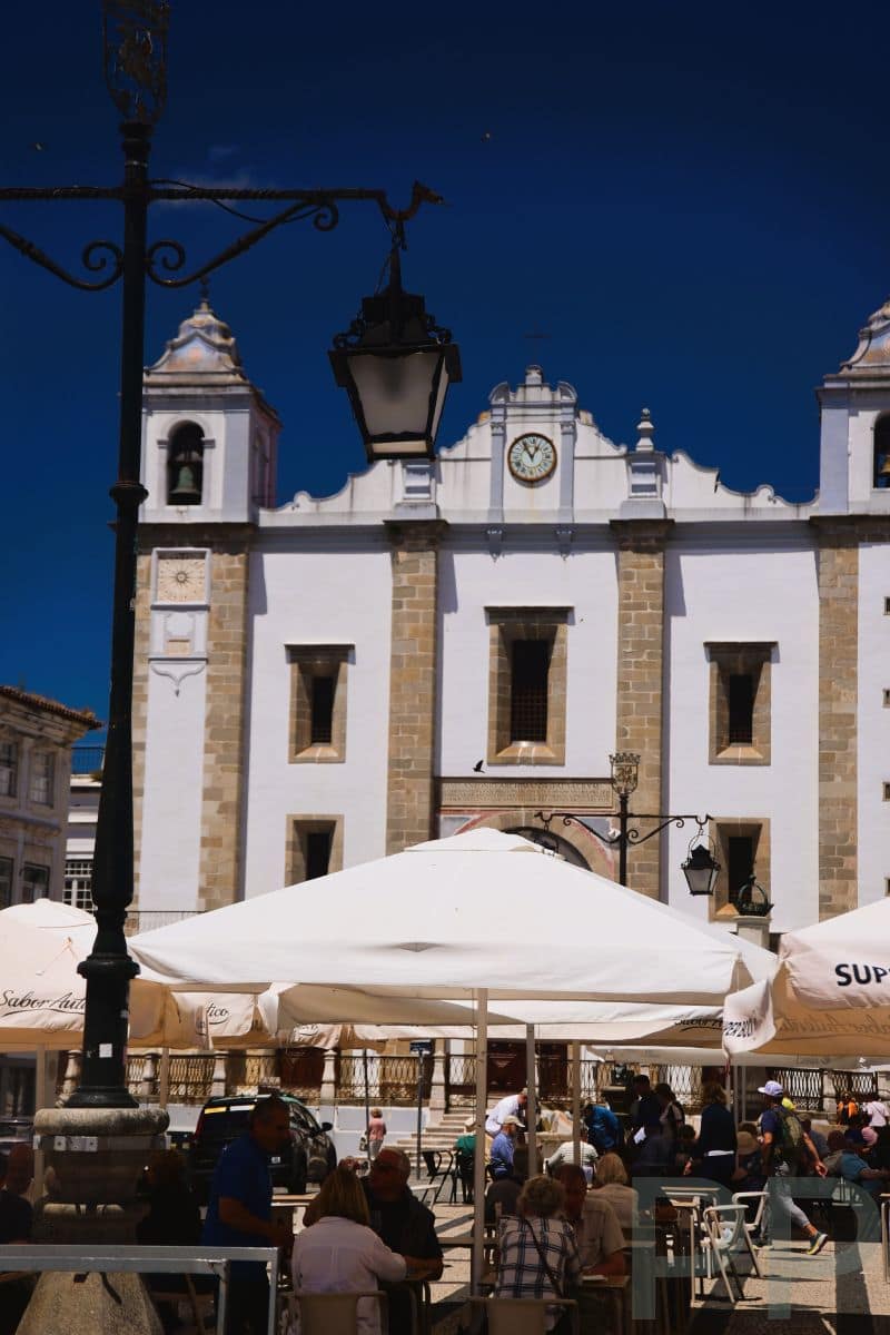 Outdoor café tables with umbrellas on Praça do Giraldo in Évora, Portugal, with the Church of St. Anthony in the background.