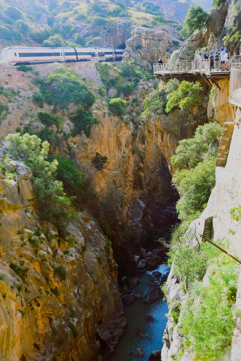 Train emerging from a tunnel above the Guadalhorce River with hikers on the Caminito del Rey boardwalk across the gorge.