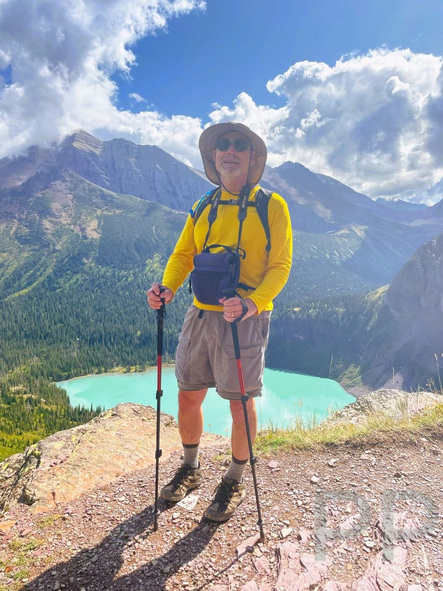 Hiker with trekking poles standing on a rocky trail overlooking turquoise alpine lake in Glacier National Park