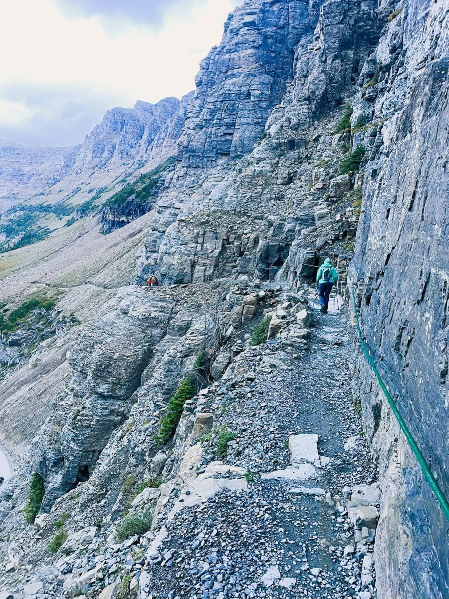 Hiker on the Highline Trail ledge section above the Going-to-the-Sun Road with green safety ropes.