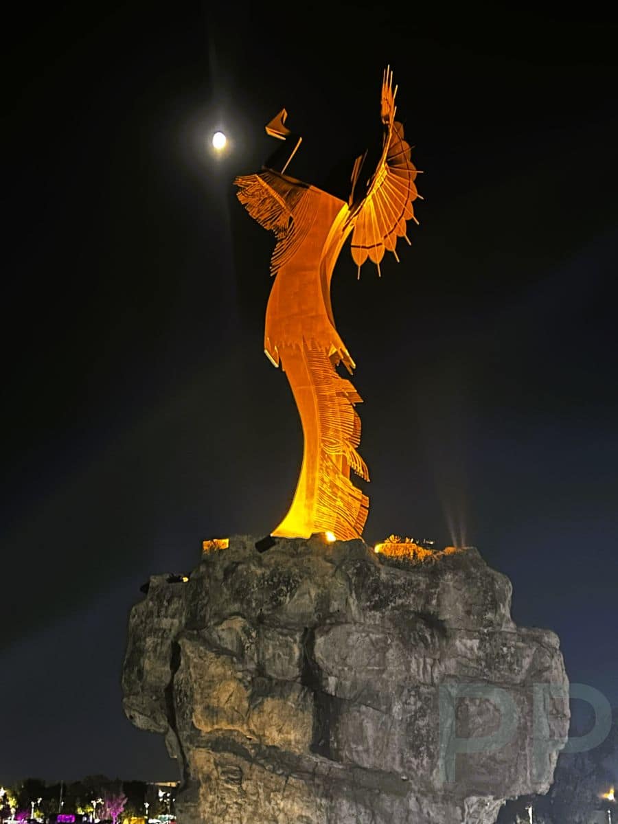 Close-up view of the Keeper of the Plains lit at night with the moon shining behind the statue.