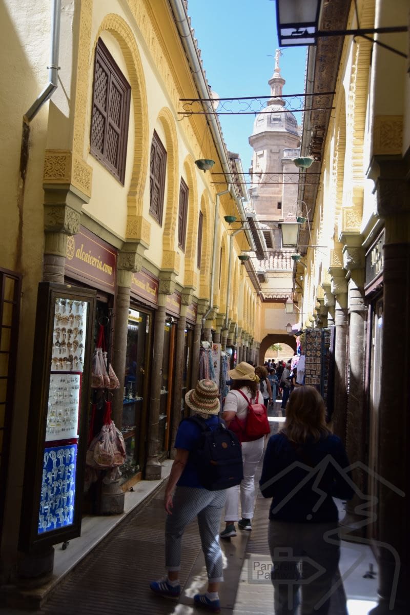 Narrow market street in Granada&rsquo;s Alcaicer&iacute;a with shop displays and hanging signs.